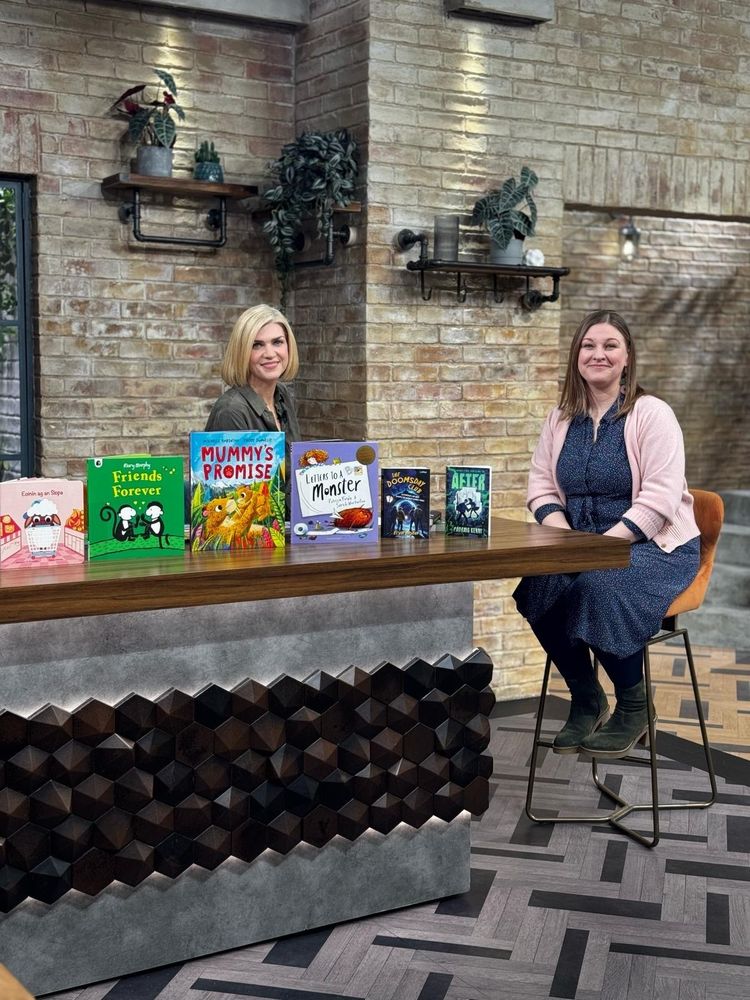 Elaina and Muireann both sit at a large table on the set of Ireland AM. They are sitting on high stools and on the dark brown wooden table in the foreground are 5 Children's books. Both Elaina and Muireann are looking at the camera and smiling.