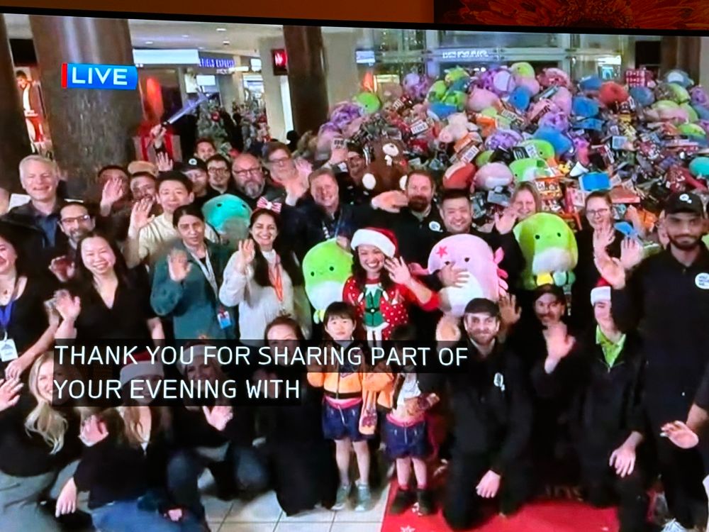 Photo of a television screen and a broadcast of a big group of people standing in front of a giant pile of toys. They’re all waving and closed captioning on screen says THANK YOU FOR SHARING PART OF YOUR EVENING WITH…