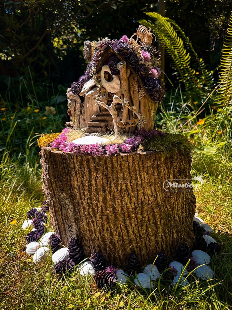 A stump on some grass in the sunshine with white shells and purple pine cones circling its base. On top of the stump, some moss and flowers, and a fairy house made of sticks and pine cones