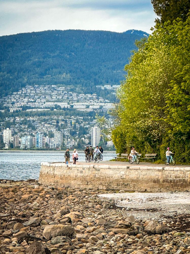 People walking and cycling on the paved Seawall path that borders Stanley Park. This photo is taken from a distance across a rocky beach at low tide. There is water to the left and in the background of the photo. Across the water is the skyline of West Vancouver with apartment towers and houses leading up the mountain, where they meet the treeline.