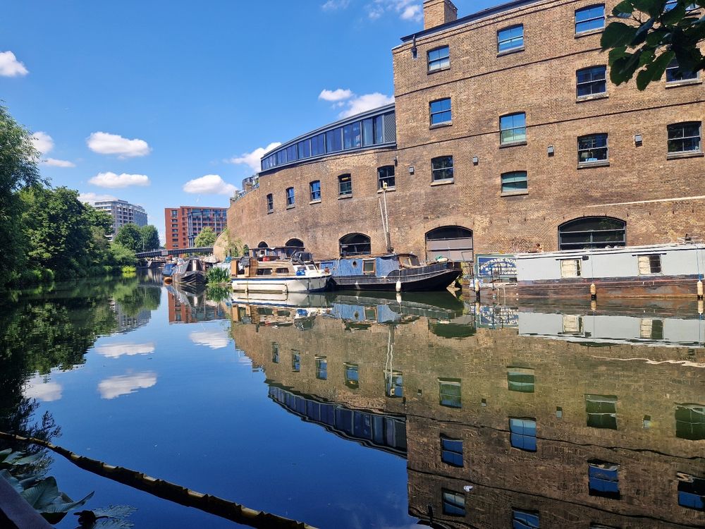 View of Regent's Canal from Camley Street Park. Still water,reflections of sky and old warehouse buildings. Narrowboats along the towpath. Not a balaclava in sight...