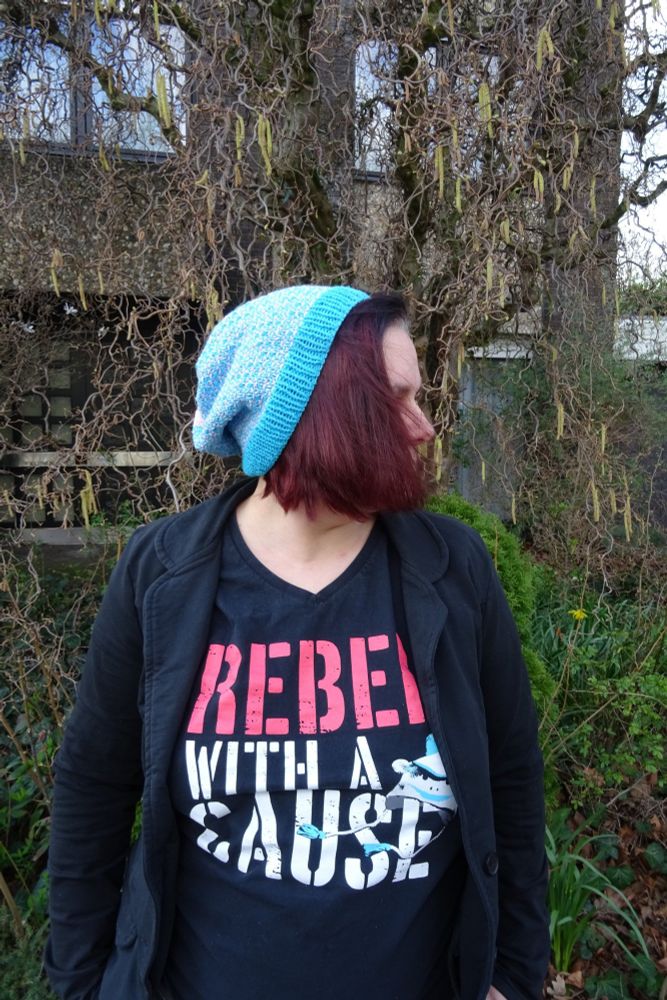 Me, a white woman with reddish shoulder long hair, dressed in black with a shirt that says "Rebel with a cause". I have my head turned away from the camera, on my head is a blue and pink handknit slouchy beanie. The brim is blue, followed by pink and blue mixed in linen stitch, the crown is solid pink