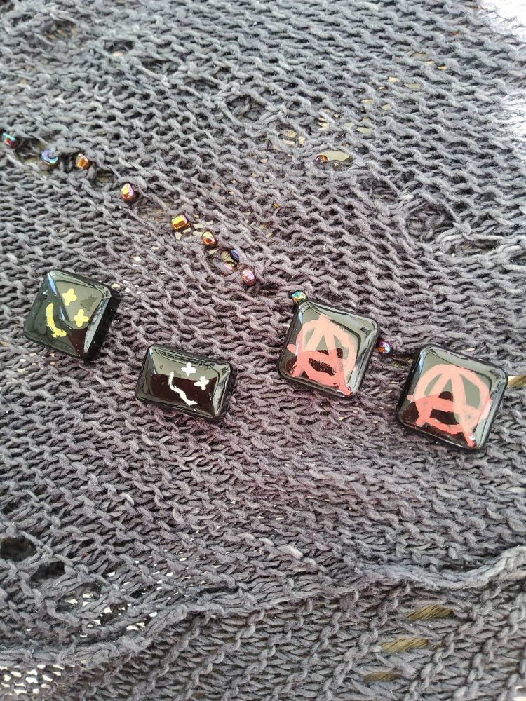 on a grey knit shawl with iridescent beads sit 3 square and one rectangular pieces if black resin with bright patterns.  Square on the left has a yellow smiley face with x-eyes, next us a rectangle with a white smiley face, on the right two squares with red anarchy signs.