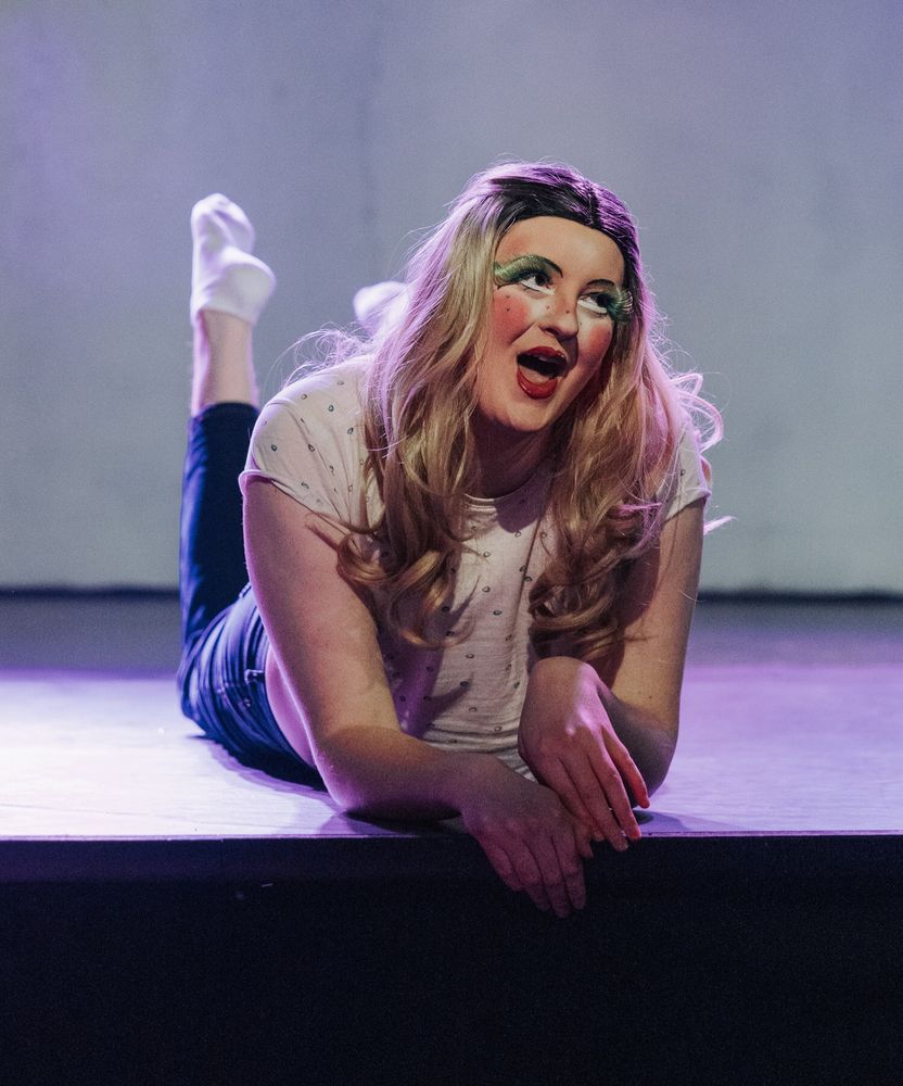 Cass Cumulus as drag queen Cassandra lays on stage on her belly with her face up looking happy and excited. Her face has exaggerated doll-like feminine features with pink brush, pink-red big lips and green fluttery fake eyelashes. She also has little hearts in different colors drawn like freckled on her cheeks and nose. She is wearing a long blonde wig, pink crop top with little hearts and high waist jeans. Her feet are up behind her.