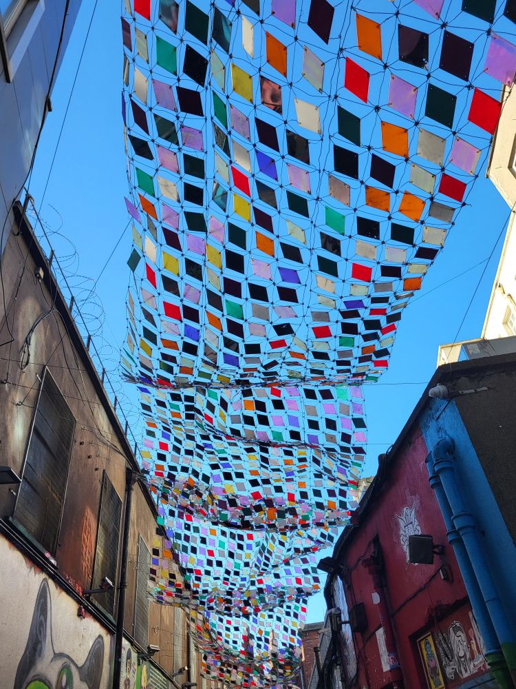 Über einer schmalen Gasse hängt eine Art Teppich aus bunten Kunststofffliesen in einem Netz