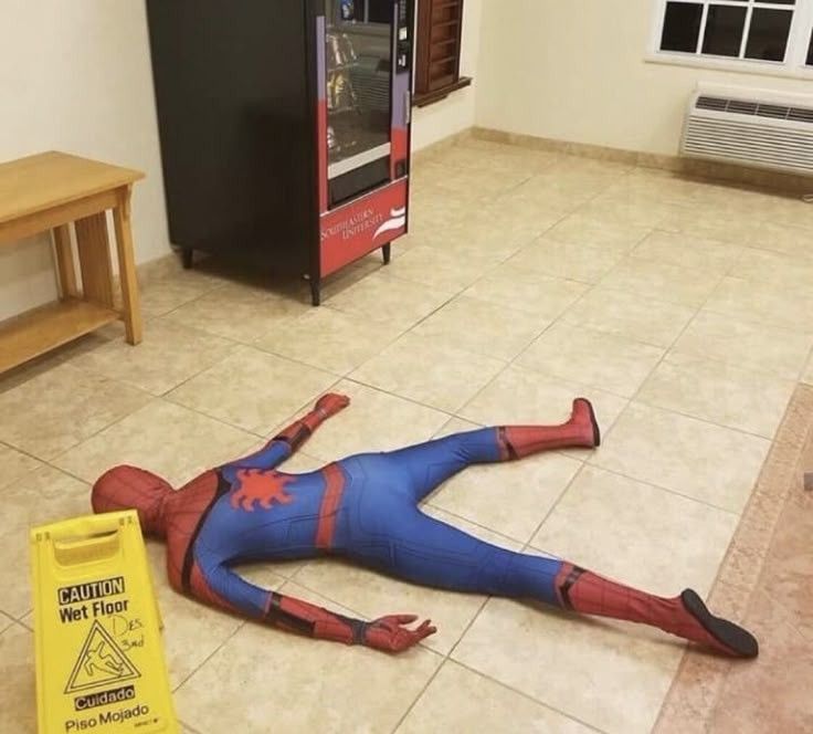 Homem-Aranha caído estrupiado no chão ao lado de uma placa dizendo que o chão está molhado 