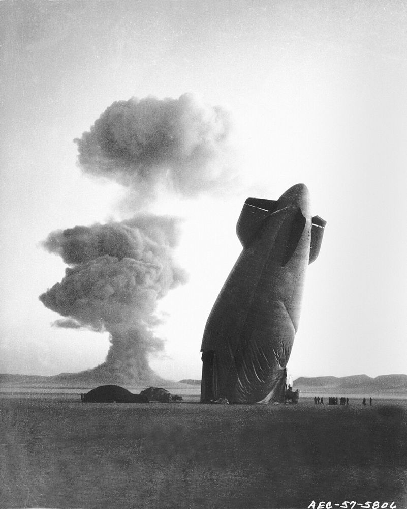 Black-and-white photo of the 1957 Operation Plumbbob nuclear test, showing a towering mushroom cloud rising in the distance while a large, partially deflated blimp leans dramatically to one side in the foreground, with small human figures visible nearby.