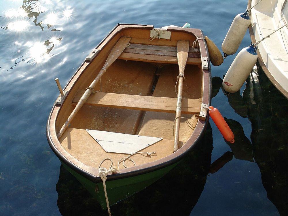 A simple wooden rowboat tied up next to a larger vessel, with two oars lying across benches with room for maybe 4 people.
