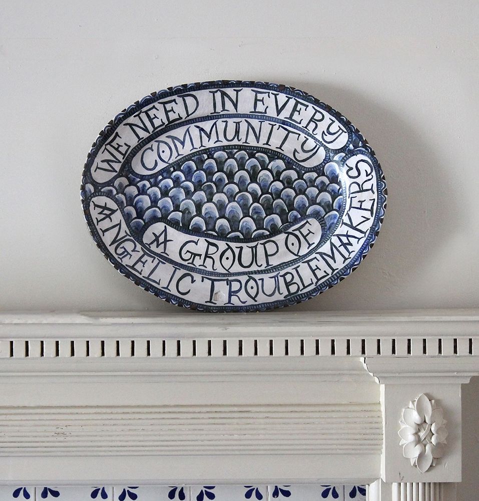 Photograph of an oval blue and white platter on an ornate white mantelpiece. The platter is decorated with a scale pattern and the text, by Bayard Rustin, reads ‘we need in every community a group of angelic troublemakers’.