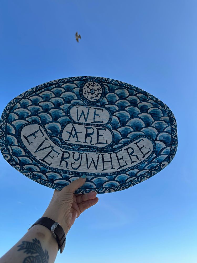 A hand holding up a palm leaf platter to the sky. The platter has blue drawn decorations in a scale pattern and the words ‘we are everywhere’.