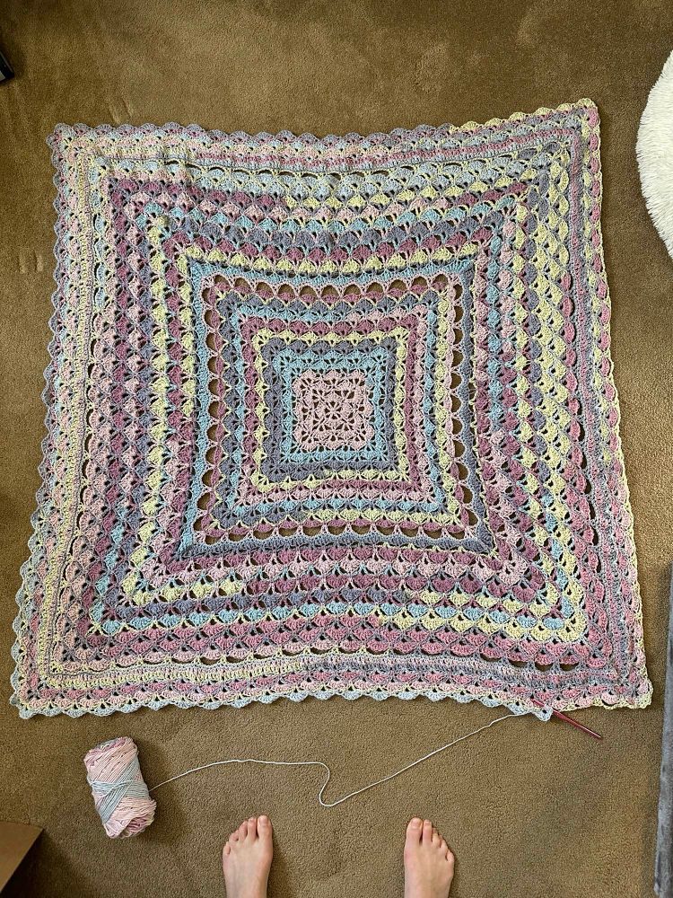 multi-color crochet blanket laid out on the floor. the blanket mostly uses the shell stitch with some solid rows and larger shell stitches ro create accents that break up the pattern