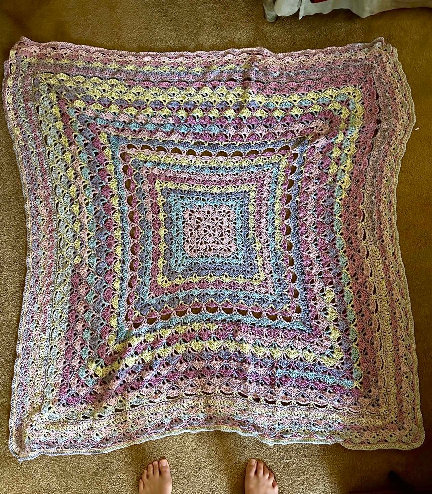 multicoloured crochet blanket laid out across the floor. the blanket is mostly made up of the shell stitch creating a loose and delicate pattern
