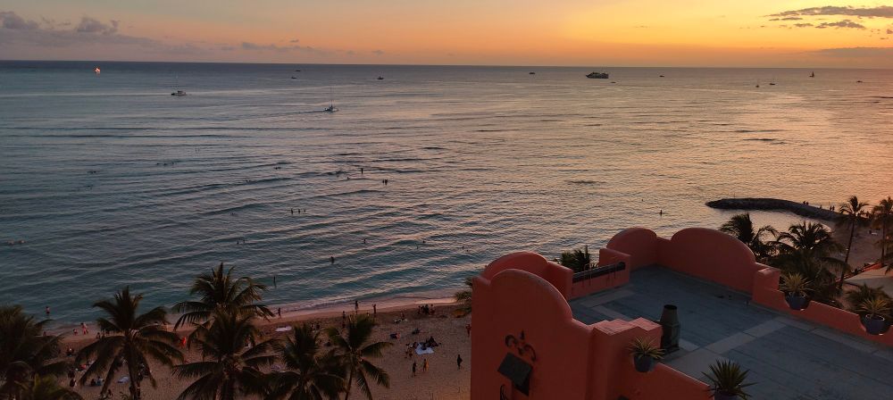 view of a Hawaiian beach at sunset from a high floor of a resort. the water is dotted by at least a dozen people, and palm trees and a pink hotel building line the shore 