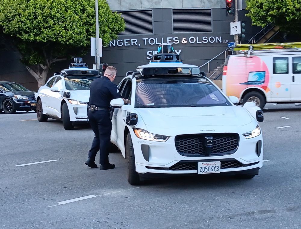 Several waymos blocking an intersection near the LA Hands Off protest while one very exasperated cop tries to make them move (they did not)