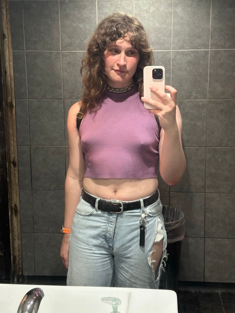 a girl stands in front of a mirror in the bathroom at a queer club event, taking a selfie. she's wearing a chain around her neck, ribbed purple crop top, jeans with a big hole on the right, a black belt, an orange watch, and a leather flag reading PAIN clipped onto her front right jeans side