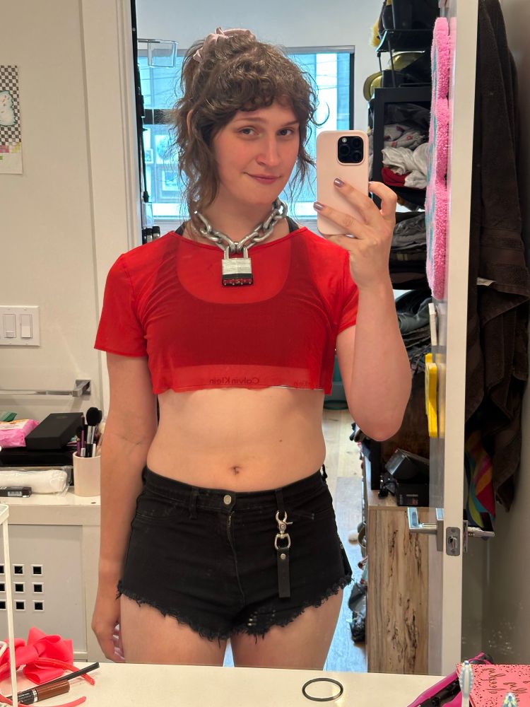 a selfie of a girl in front of a mirror, wearing a heavy metal chain and lock around her neck, a sheer red crop top over a black calvin klein bra, frayed cutoff shorts, a black leather clip flagging PAIN on the right, with her hair up in a ponytail