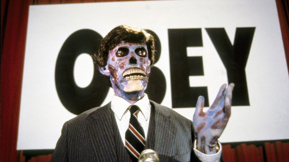 Image of skeletal figure in suit with a sign reading “obey” behind him. This image is from the 1988 film “They Live” directed by John Carpenter. 