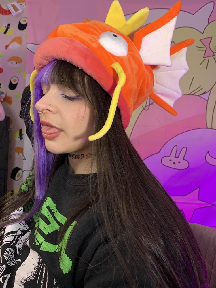Picture of Quartsy from 2023 wearing faer Magikarp Pokemon Center hat in fey's stream room. Fey had split dyed purple and brown hair at the time, and was wearing an attack on titan hoodie. Eyes were closed and sticking tongue out in a silly pose.