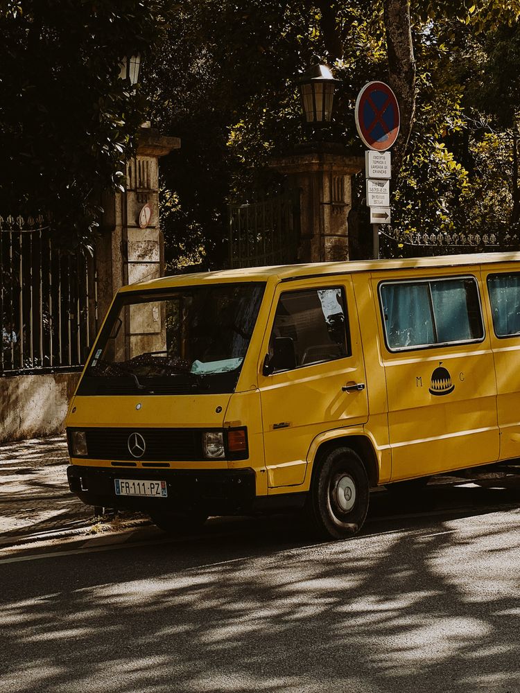 A yellow Mercedes-Benz van parked on a street next to a no-parking sign with trees in the background.