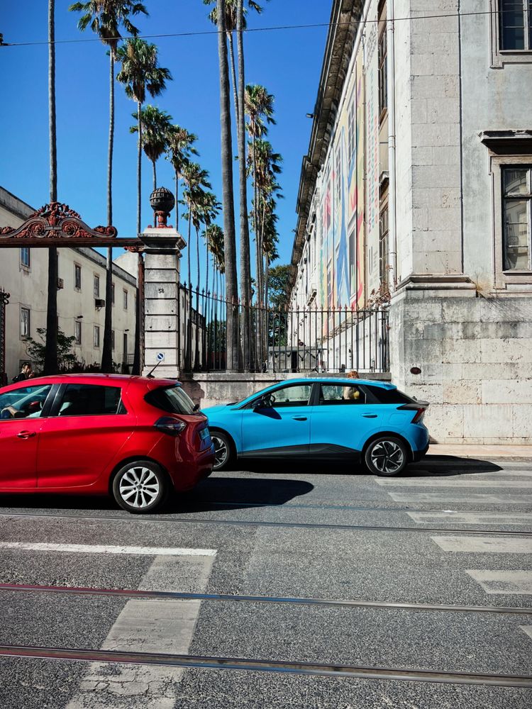 A red car drives by a blue car. Some palm trees in the background. City street. 
