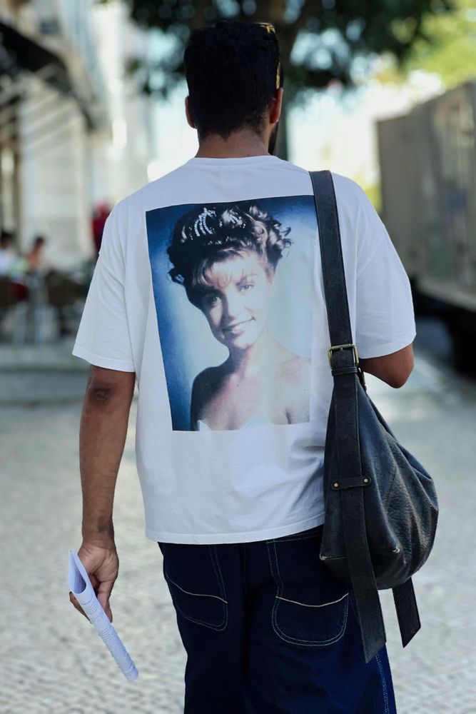 A person is walking down a cobblestone street, seen from behind. They are wearing a white T-shirt with a large printed photo of Laura Palmer. The person is carrying a black shoulder bag and holding some papers in their left hand. The background has blurred buildings and trees.