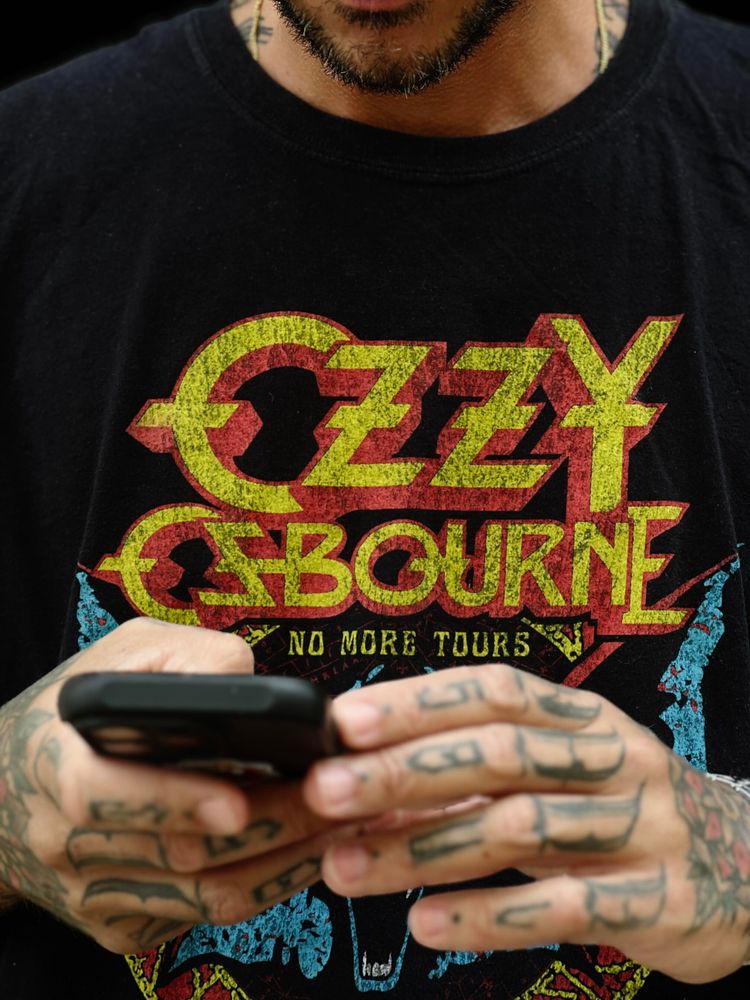 A person is holding a smartphone, focusing on the screen. They are wearing a black T-shirt with bold yellow and red lettering that reads "Ozzy Osbourne" and "No More Tours" in smaller text. Their hands and neck have visible tattoos.