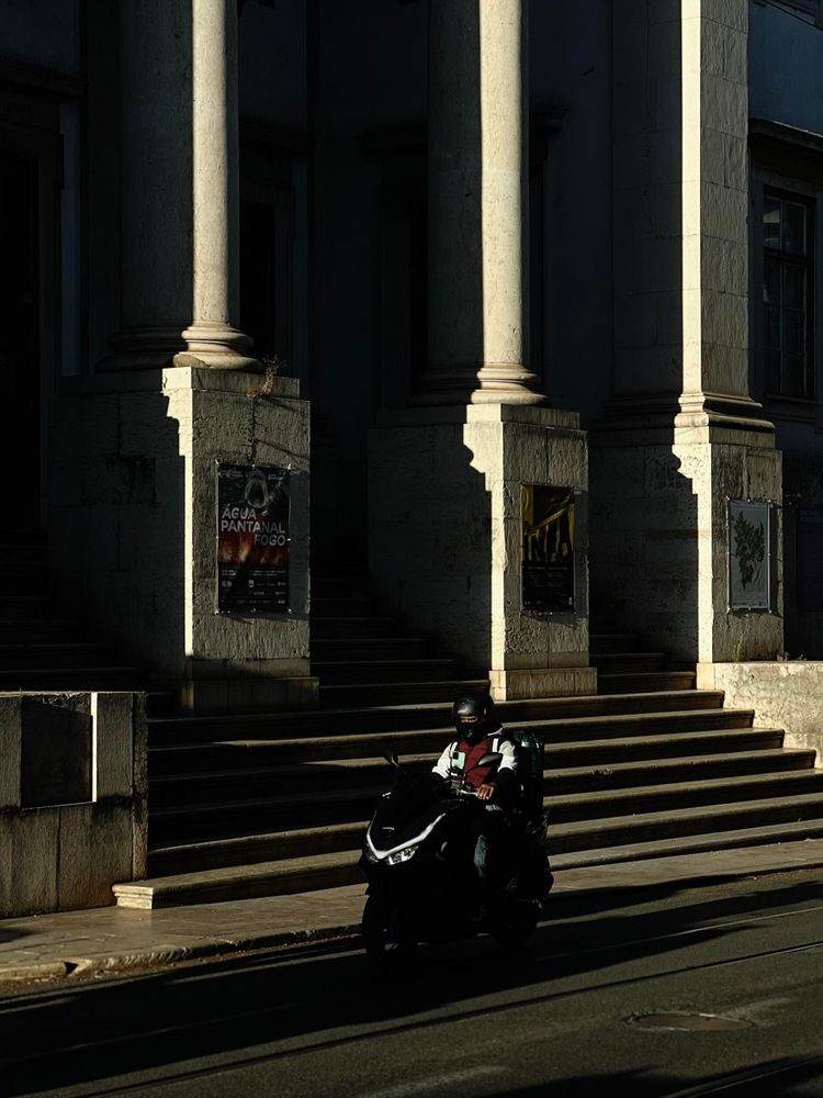 A person on a scooter rides past a building with large stone columns and steps in the background. The scene is mostly in shadow with patches of sunlight highlighting the architecture. Posters are visible on the walls near the steps. The person on the scooter is wearing a helmet and carrying a green backpack.