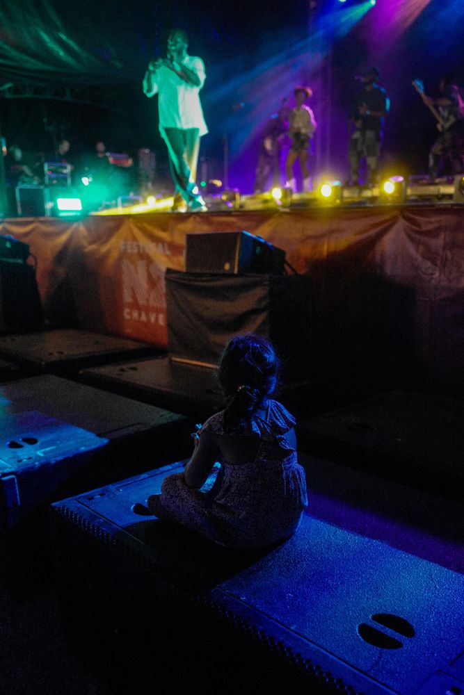 A young child sits on a speaker at an outdoor concert, facing the stage where musicians perform under colorful lights. The stage is elevated, and the child appears to be watching the show intently. The scene is illuminated with vibrant blues, purples, and yellows, creating a lively atmosphere.