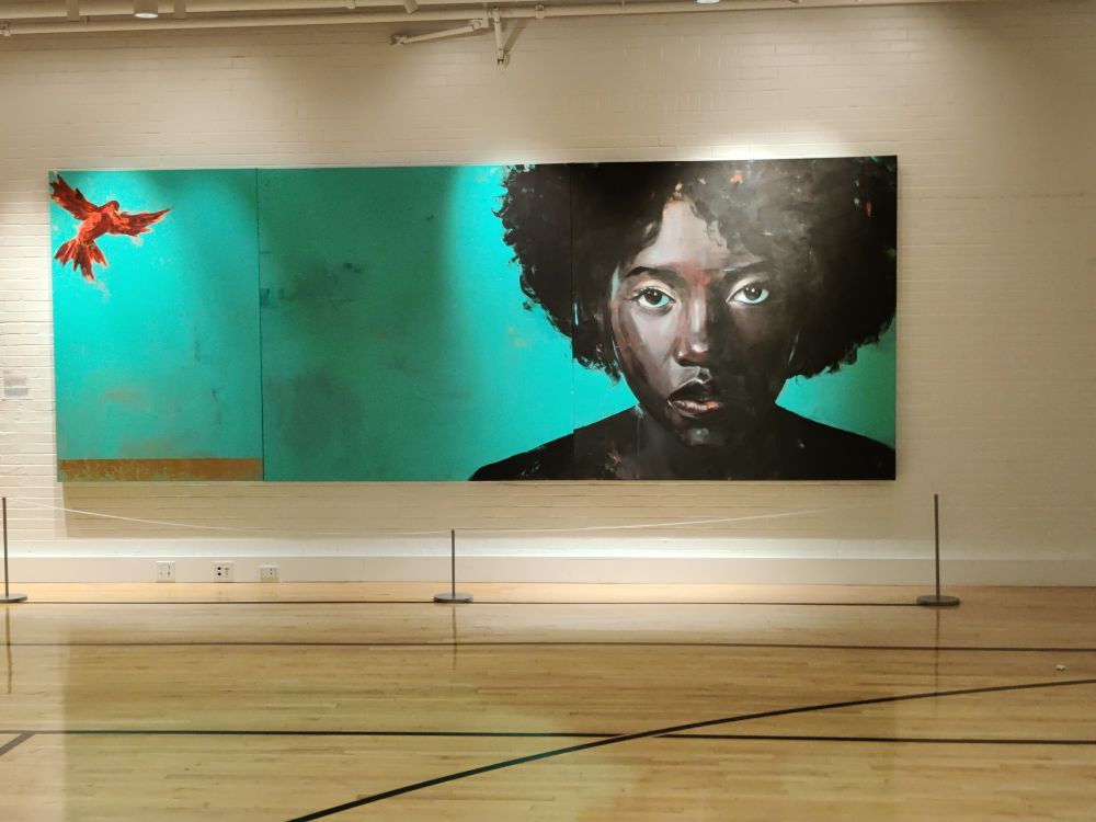 On the left side of the painting is a red birth and on the right side is a Black women from shoulders up. The background is green. The floor reminded me of a gymnasium. The bird and women have spot lights on them.