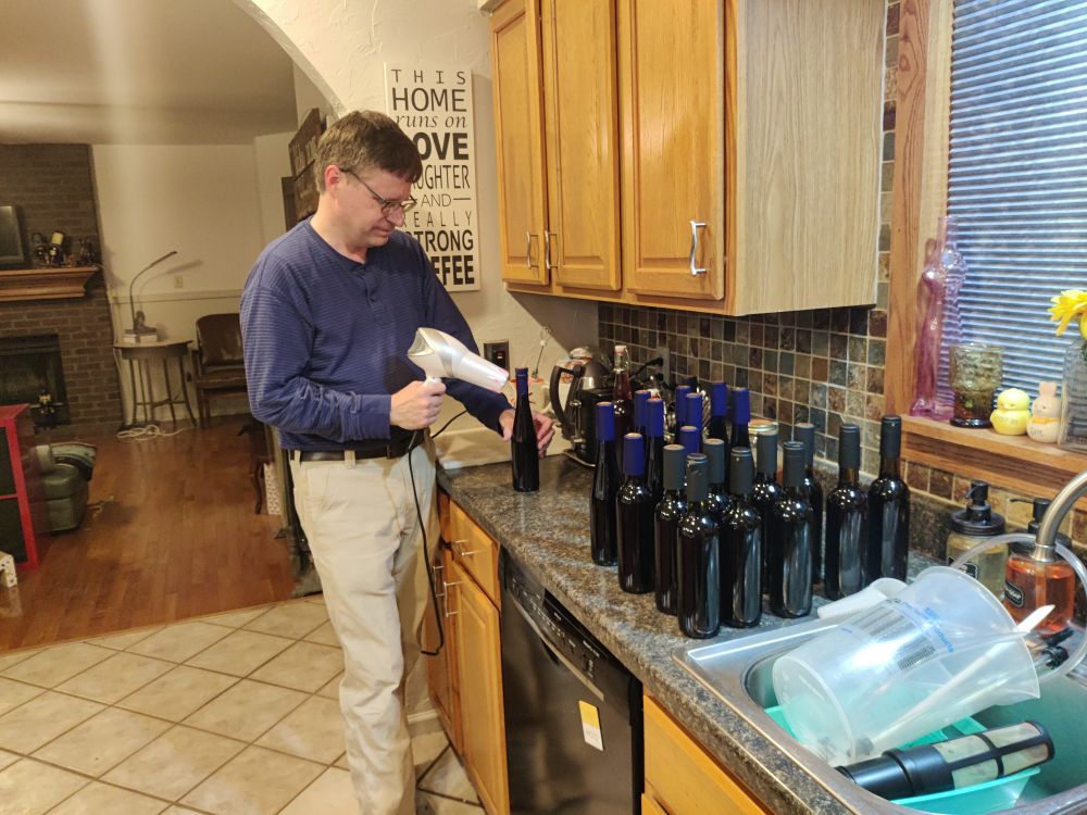 My husband holding a hair dryer in one hand and a 375 ml blue bottle in the other. About 20 bottles are in a line waiting their turn. Ignore the dirty dishes in the sink.