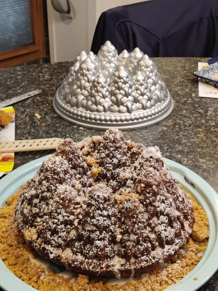 In the background there's a silver mold that looks like a ring of trees. In the foreground is a cake that used the mold. The cake is white because of a dusting of powdered sugar. The light brown ring was made with cake mix and butter, just enough butter to make it crumble. My choice to leave it in the frig overnight made it hard & difficult to work with, thus it was used around the edge of the cake, resembling a forest floor. The cake and crumble are on a green base.