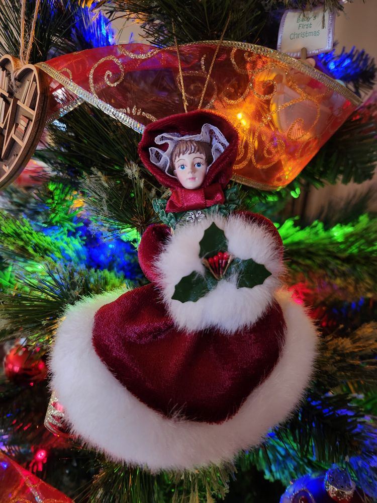 A Christmas ornament of an 1800s Carol singer in a red velvet cloak with white fur trim holding a sprig of holly