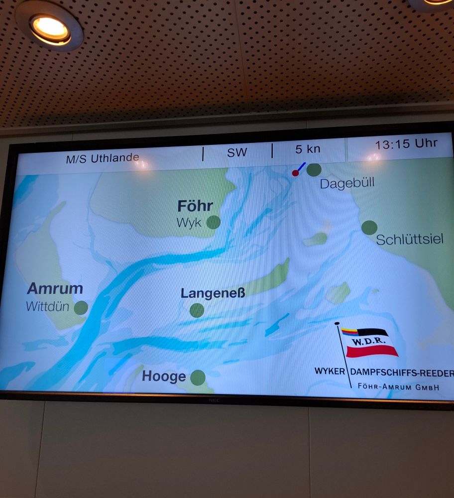 Ein Bildschirm, auf dem Dagebüll sowie die Inseln För und Amrum und die Halligen Langeneß und Hooge abgebildet sind. Ein roter Punkt kurz hinter Dagebüll zeigt den aktuellen Standort der Fähre. Rechts unten in der Ecke ist das Logo der W.D.R. 