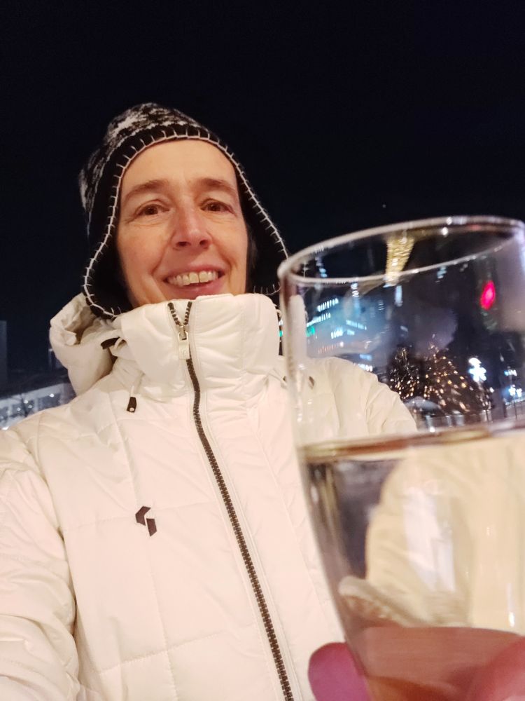 Eine Frau in weißem Anorak und einer schwarzen Mütze mit weißem Muster hält ein Sektglas in der Hand. Im Glas spiegeln sich die Lichter eines Weihnachtsbaums. Der Himmel ist dunkel. 