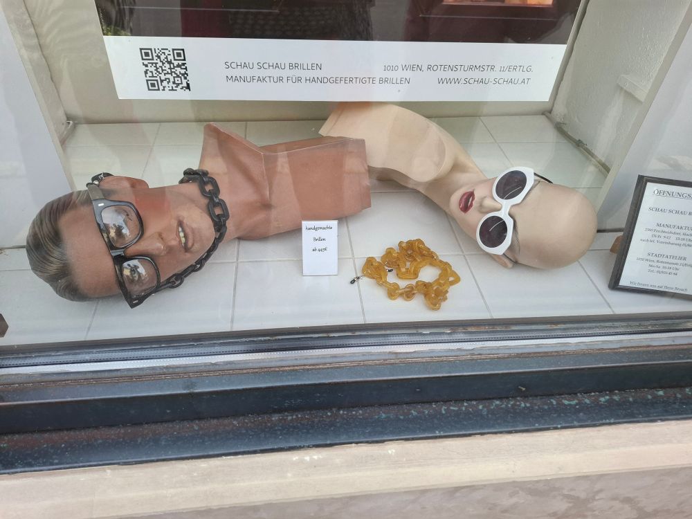Ein Schaufenster mit den Köpfen von zwei Schaufensterpuppen. Eine ist männlich, die andere weiblich. Der Männerkopf trägt eine durchsichtige Brille und eine schwarze Kette um den Hals. Der Frauenkopf (ohne Haare) trägt eine Sonnenbrille. Zwischen den Köpfen liegt eine Bernsteinkette.
