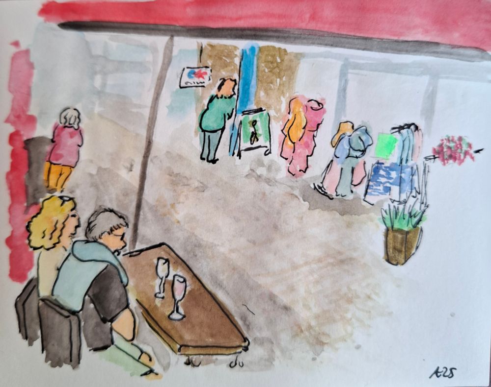 Zwei Personen, ein Mann und eine Frau, sitzen an einem Tisch. Auf dem Tisch stehen zwei Weingläser. Links steht noch eine Frau, eine weitere gegenüber, bei einem EC Automaten. Gegenüber dem Tisch, auf der anderen Straßenseite, steht ein Kübel mit einer Grünpflanze. Dahinter hängen Kleider auf einem Ständer und auf dem Boden steht ein Schild mit Werbung. 