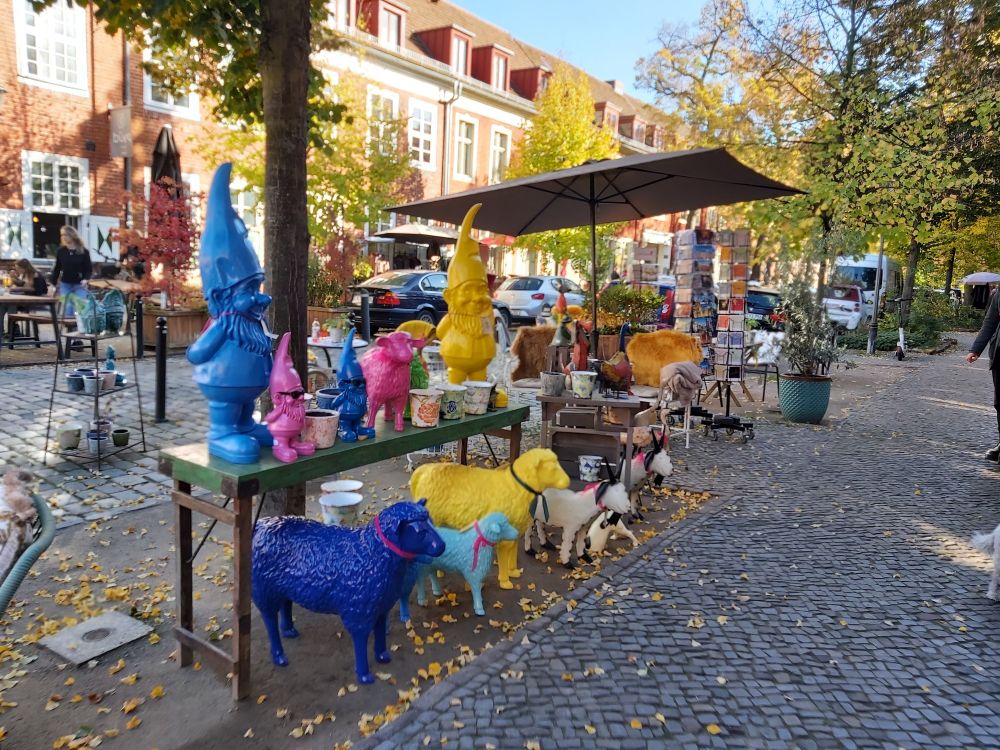 An einer gepflasterten Straße stehen auf einem Tisch bunte Gartenzwerge. Sie sind einfarbig in blau, pink und gelb. Unter dem Tisch stehen ebenso bunte Schafe. 