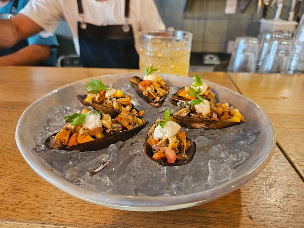 Prato cinza de petisco, sobre um balcão de bar marrom, o prato tem gelo dentro, por cima do gelo temos 5 metades de Ostras preenchidas com pedaços de mariscos e vinagrete. Atrqs do prato temos um copo de vidro com um drink laranja. Atrás do balcão é visto sem destaque o garçom preparando outras bebidas.