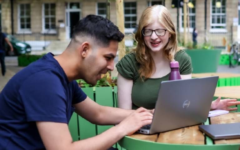Close-up photo of two differently-looking students sitting at a round table looking at a laptop outdoors in the quad of UCL with the main building behind