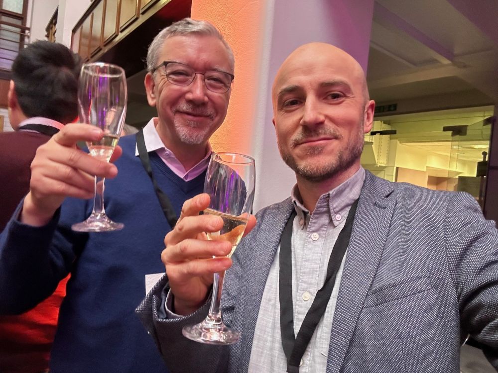 Two men, each holding a glass partly full of wine or champagne, standing together in a modern, partitioned room with other people and offices in the background.  They are smiling, dress smartly and wearing their passes on black ribbons hanging around their necks