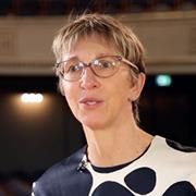 A colour photo of Barbara Conradt, from sternum to top of head, wearing a navy-large-spotted white dress and curved, light brown-framed spectacles, giving a presentation (not visible).
