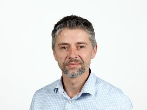 Headshot of man having grey-dark, short hair, beige face, brown eyes, close-cut grey-brown beard and moustache, wearing a pale blue and white small-checked shirt with an open collar