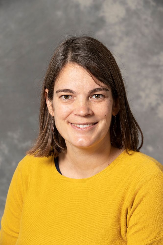 A head-and-shoulders photo of a smiling Anna Franz, a woman having mid-length, straightish, side-parted brown hair and brown eyes, wearing a bright mid-yellow jumper and silver dangly earrings 