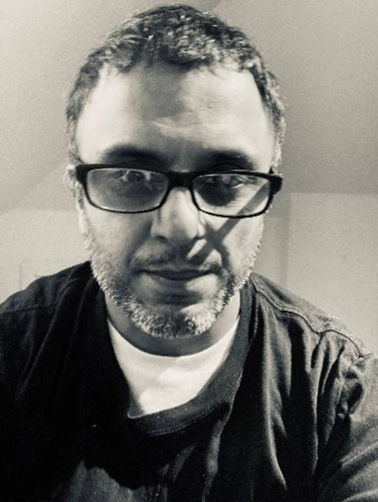 A black & white photo of Sandip Patel from sternum to head, wearing a white T shirt with a darker top over it and dark-rimmed, narrow rectangular glasses.