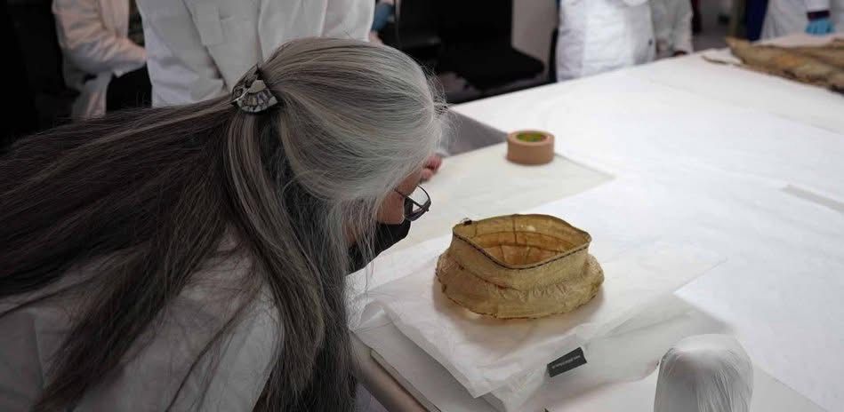A woman with long grey hair and a mask looking at a traditional belonging of bark or fabric 