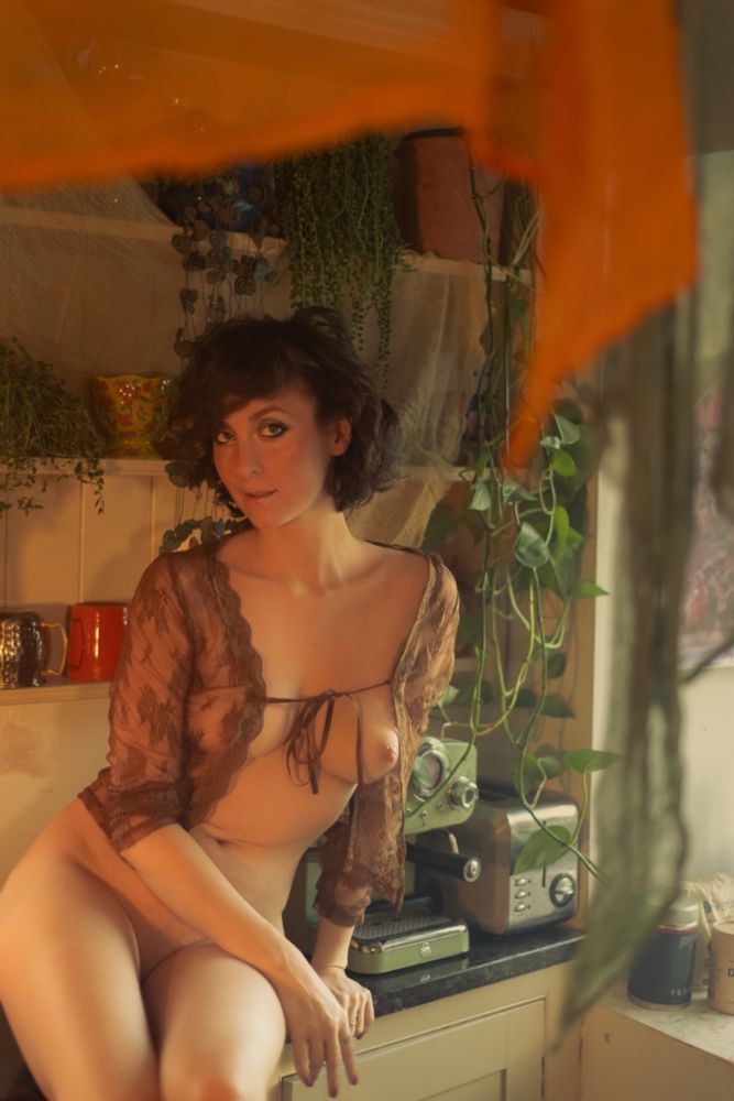 Naked female with a sheer top sitting on a kitchen bench with breasts on show