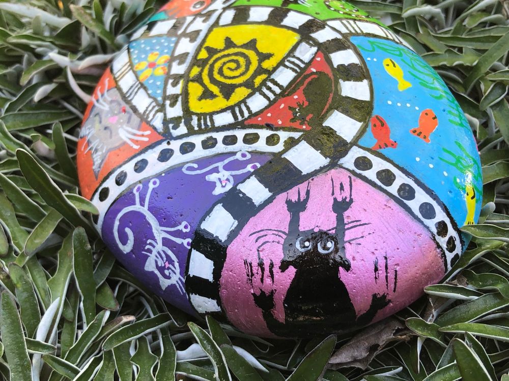 Painted rock patchwork style with black white ribbons separating different colored backgrounds.  Inside sections are black cat sliding down wall, sun, fish, white skeletal cat, grey cat face.