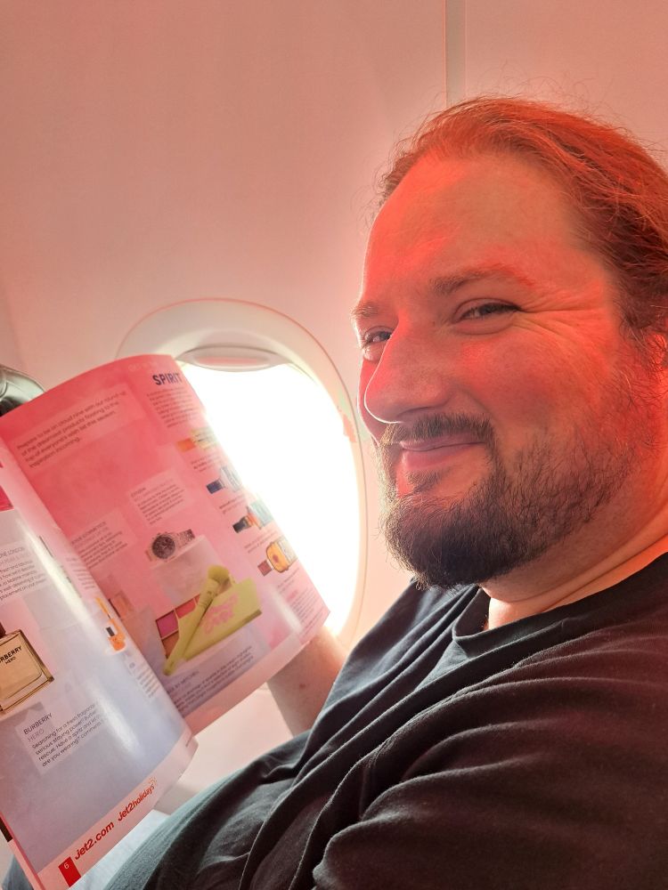Antony, a white person with a beard, smiles at the camera while reading the in-flight magazine on an aeroplane. 