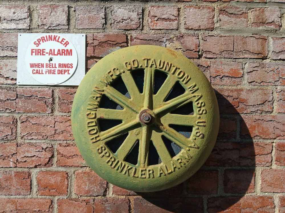 An old green metal wheel that says "Sprinkler Alarm"- next to the modern white&red sign, "Sprinkler fire-alarm"