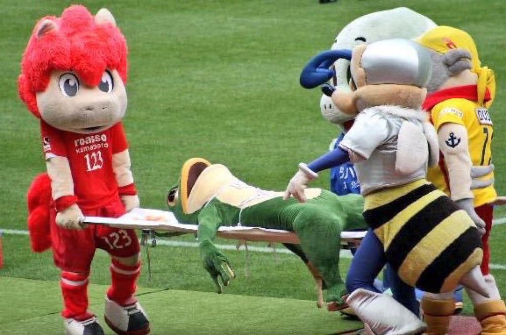 Someone dressed as a frog mascot for a sports team is on a stretcher being carried by other mascots. His mouth is agape and eyes staring out. He may be dead, or wishes he were. No one has suffered as much as him.